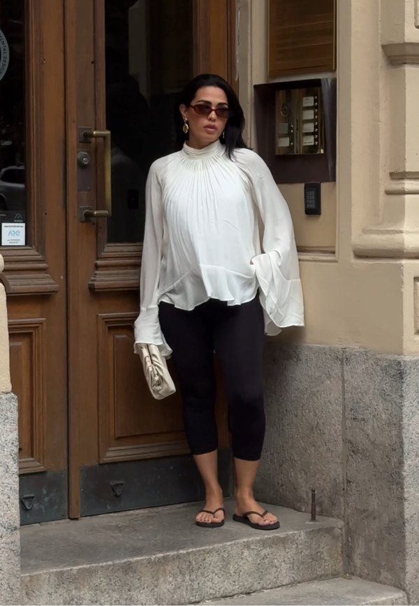 Femme portant un chemisier blanc ample, des leggings noirs courts, des tongs, des lunettes de soleil, tenant une pochette beige, debout près d'une porte en bois.