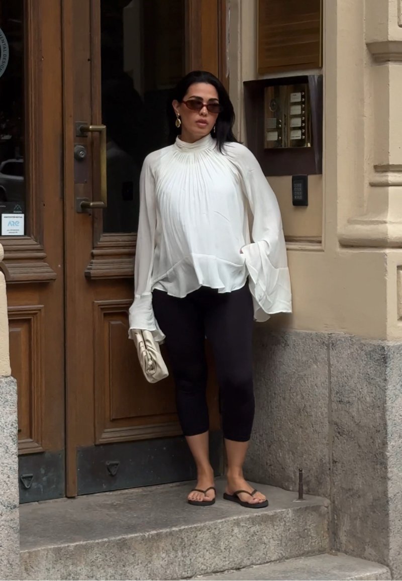 Femme portant un chemisier blanc ample, des leggings noirs courts, des tongs, des lunettes de soleil, tenant une pochette beige, debout près d'une porte en bois.