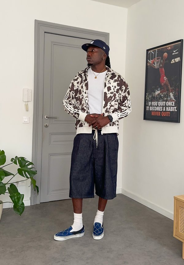Mand står indendørs iført en jakke med ko-print, hvid T-shirt, brede denimshorts, blå kasket, blå slip-on sko og hvide sokker. Basketplakat bagved.