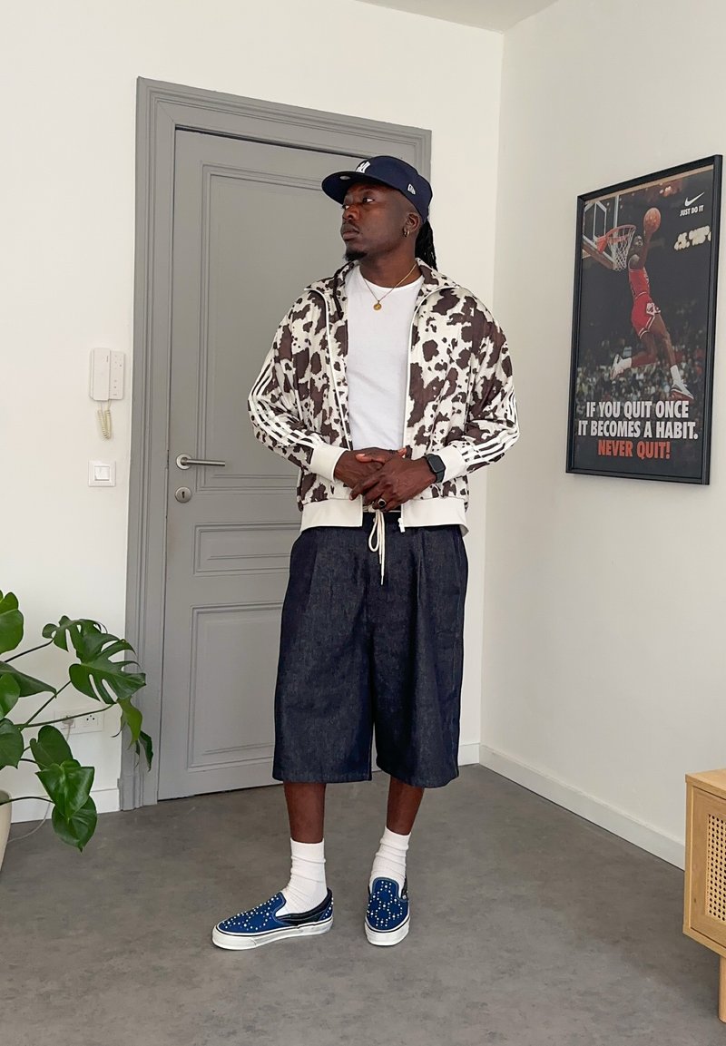 Uomo in piedi al chiuso indossa una giacca a stampa mucca, una maglietta bianca, ampi pantaloncini di jeans, un cappellino blu, scarpe blu senza lacci e calzini bianchi. Poster di basket dietro.