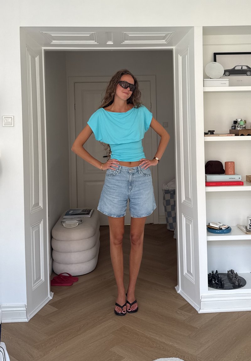 Kvinne i en turkis topp, denimshorts og svarte sandaler står med hendene på hoftene i et hvitt rom med hyller og tregulv.