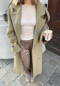Personne portant un manteau beige, un haut blanc transparent, un pantalon marron et des baskets, tenant une tasse de café, debout sur un pavé en pierre près d'une porte en bois.