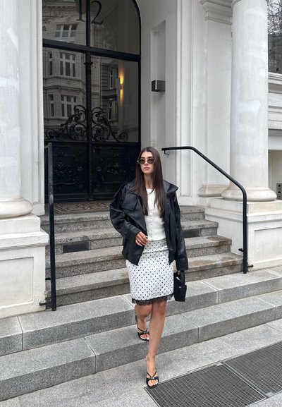 Mujer con chaqueta de cuero negra, falda blanca de lunares y gafas de sol bajando unas escaleras de piedra delante de un edificio con grandes puertas negras.