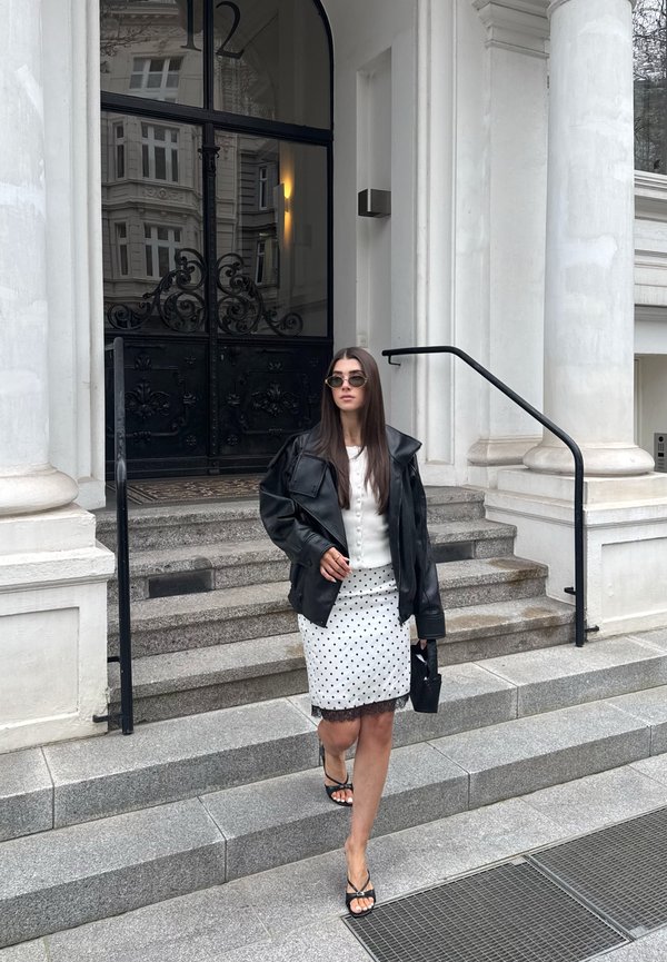 Mujer con chaqueta de cuero negra, falda blanca de lunares y gafas de sol bajando unas escaleras de piedra delante de un edificio con grandes puertas negras.