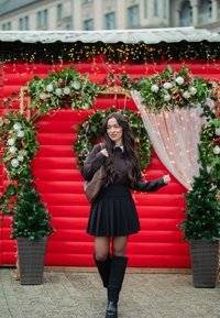 Jupe plissée noire et pull marron, assortis à des chaussettes côtelées montant jusqu'aux genoux et des bottes noires, devant un fond rouge décoré de feuillage.