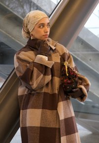 Bruin- en beige geruit wollen jas met een hoge kraag, gecombineerd met bruine leren handschoenen en een patroon tas, tegen een moderne glazen achtergrond.