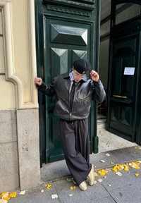 Μαύρη δερμάτινη μπomber ζακέτα, γκρι πουκάμισο και φαρδιά μαύρα παντελόνια. Στέκεται δίπλα σε μια σκούρα πράσινη πόρτα, με κίτρινες φύλλοδες στο έδαφος.