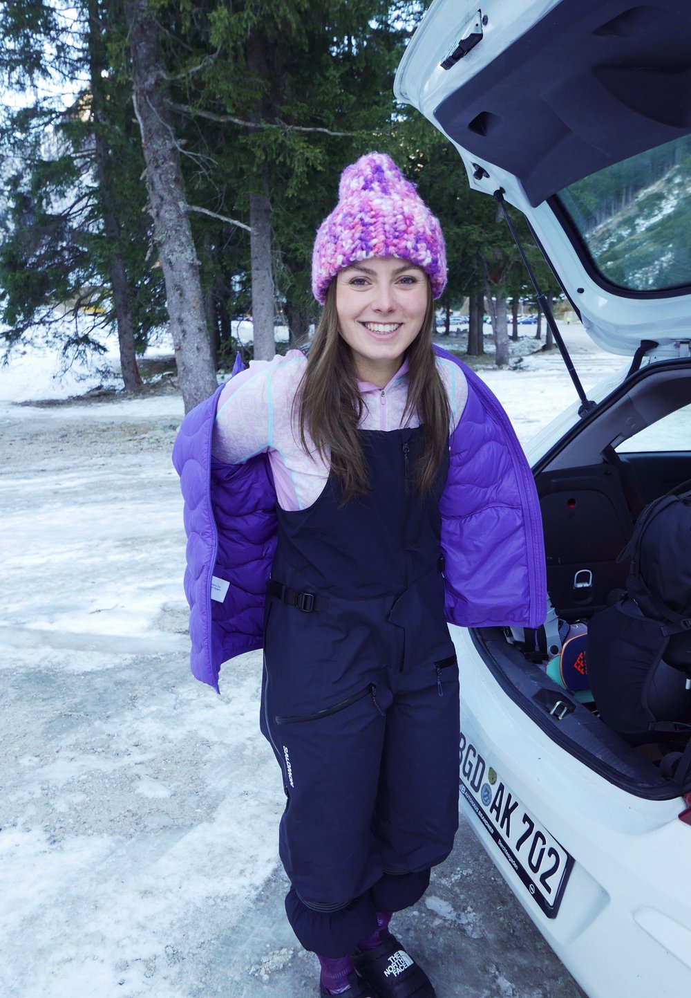 Svarte skibukser med en matchende lilla puffjakke, en rosa strikket lue og mørke sko, satt mot en snødekt bakgrunn.