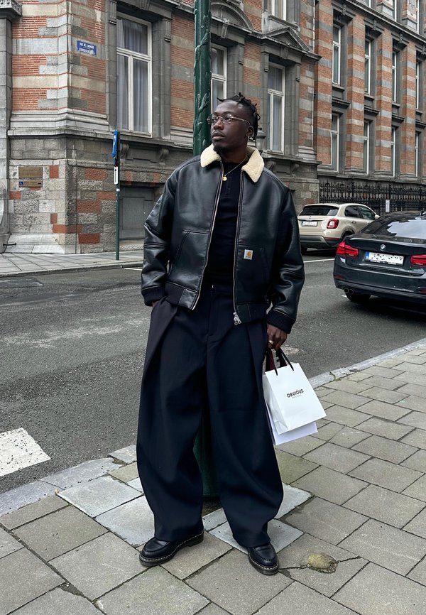 Mężczyzna w czarnej skórzanej kurtce z kołnierzem z baranka i szerokich czarnych spodniach stoi na miejskim chodniku, trzymając białą torbę na zakupy.