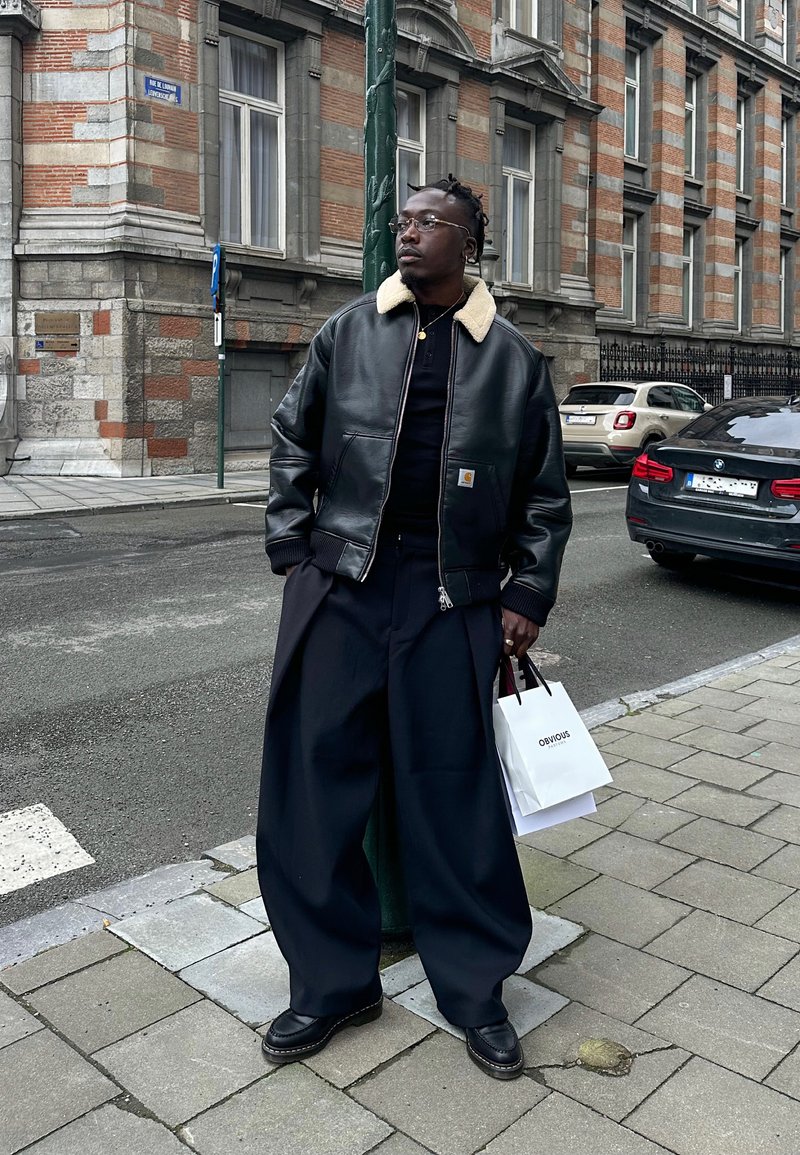 Čovjek u crnoj kožnoj jakni s ovčjim ovratnikom i širokim crnim hlačama stoji na gradskoj pločniku držeći bijelu vrećicu za kupovinu.