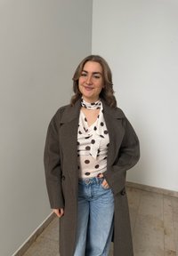 Jeune femme souriante, portant un long manteau marron sur un haut blanc à pois, avec un jean bleu clair, debout à l'intérieur contre un mur blanc.