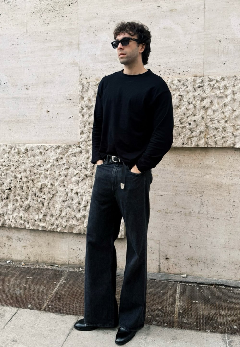 Maglietta nera a maniche lunghe, jeans larghi scuri, scarpe nere eleganti e occhiali da sole. In piedi contro un muro di pietra chiara e texture.