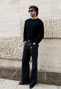 Maglietta nera a maniche lunghe, jeans larghi scuri, scarpe nere eleganti e occhiali da sole. In piedi contro un muro di pietra chiara e texture.