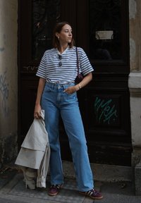 Cămașă cu mânecă scurtă în dungi, albă și bleumarin, asociată cu blugi deschiși la culoare, cu talie înaltă. Ține în mână o jachetă bej și poartă adidași violeți.