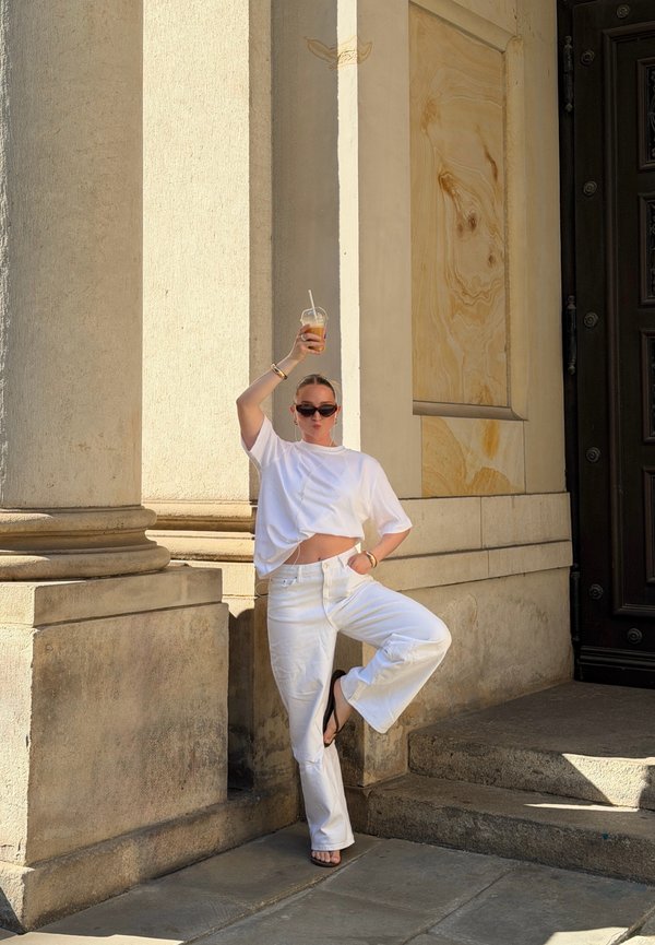 Femme vêtue de blanc et portant des lunettes de soleil, debout sur une jambe, tenant un verre au-dessus de sa tête, à côté de grandes colonnes en pierre et d'une porte sombre.