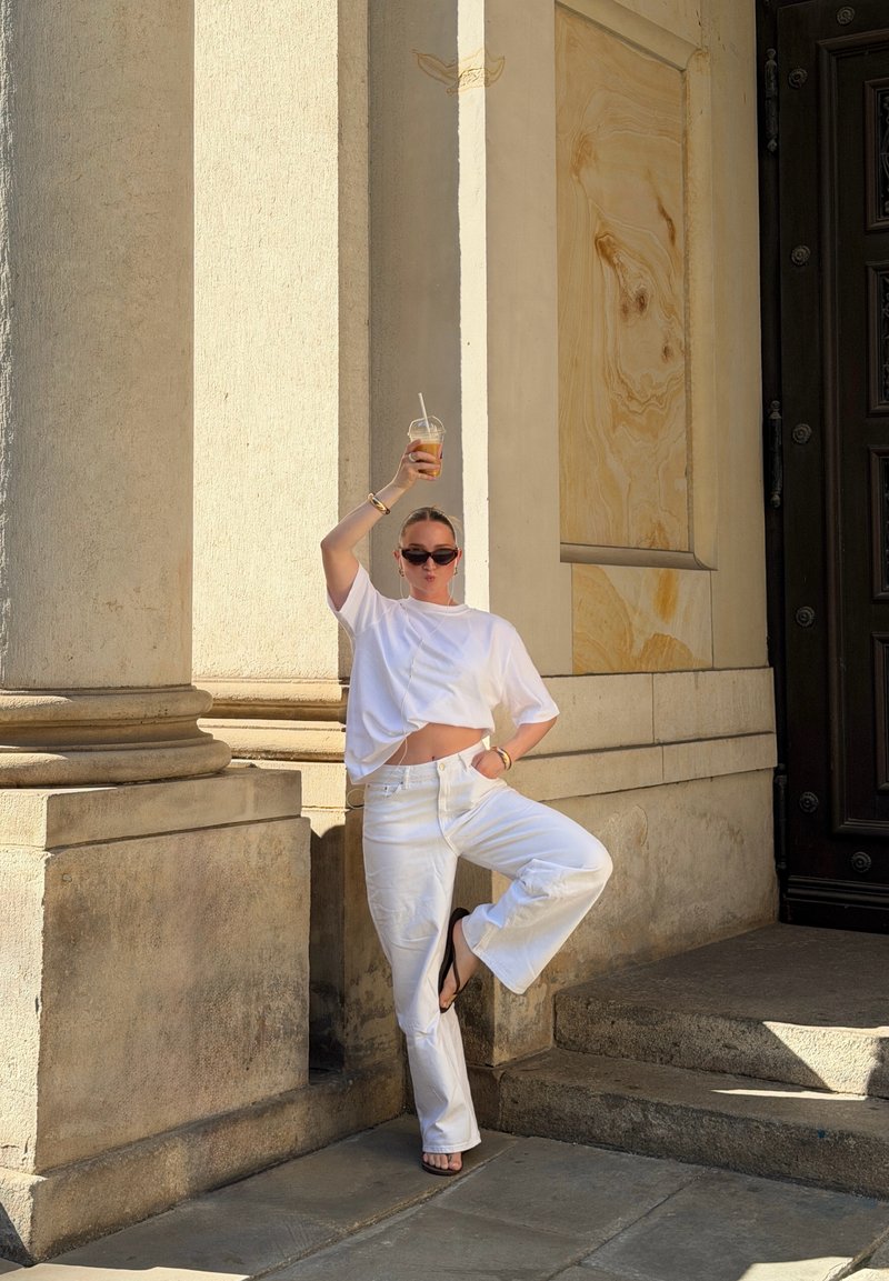 Femme vêtue de blanc et portant des lunettes de soleil, debout sur une jambe, tenant un verre au-dessus de sa tête, à côté de grandes colonnes en pierre et une porte sombre.