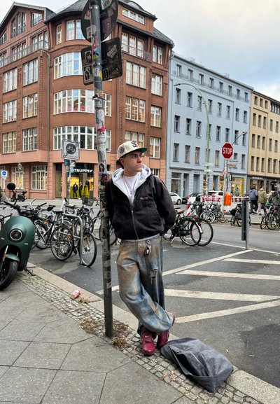 Persona apoyada contra un poste, vistiendo una sudadera negra y una camiseta blanca, y unos vaqueros claros desgastados, con una bolsa gris en el suelo.