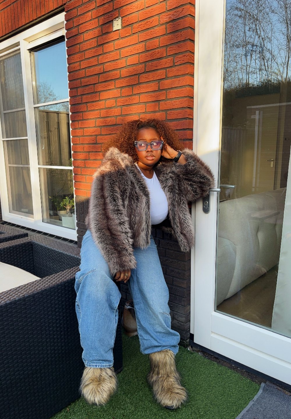 Veste courte en fausse fourrure aux tons gris, T-shirt blanc, jean bleu surdimensionné et bottes fourrées. Posée contre un mur de briques près d'une porte en verre.