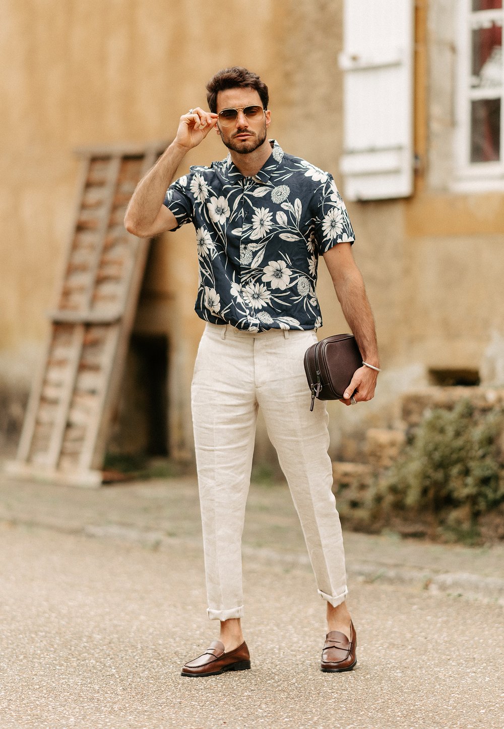 Kortärmad marinblå blommig skjorta, beige linbyxor, bruna loafers, håller en mörkbrun väska, utomhus med en texturerad vägg som bakgrund.