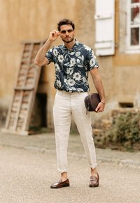 Kortärmad marinblå blommig skjorta, beige linbyxor, bruna loafers, håller en mörkbrun väska, utomhus med en texturerad vägg som bakgrund.
