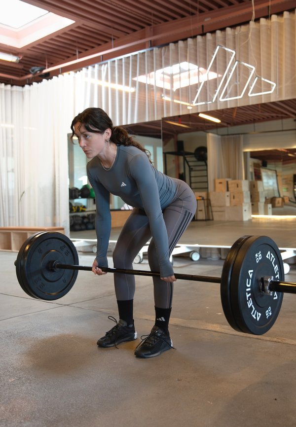 Hallis nähtav naine hallis spordiriietuses tõstab kangi, seinad on peegeldavate pindadega, treeningsaalis on treeningseadmed ja puitlaetalad.