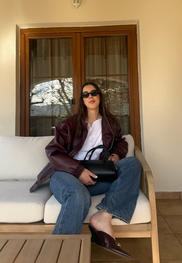 Femme portant des lunettes de soleil, une veste bordeaux, une chemise blanche et un jean, assise pieds nus sur un canapé de patio, tenant un sac à main noir devant une porte en bois.