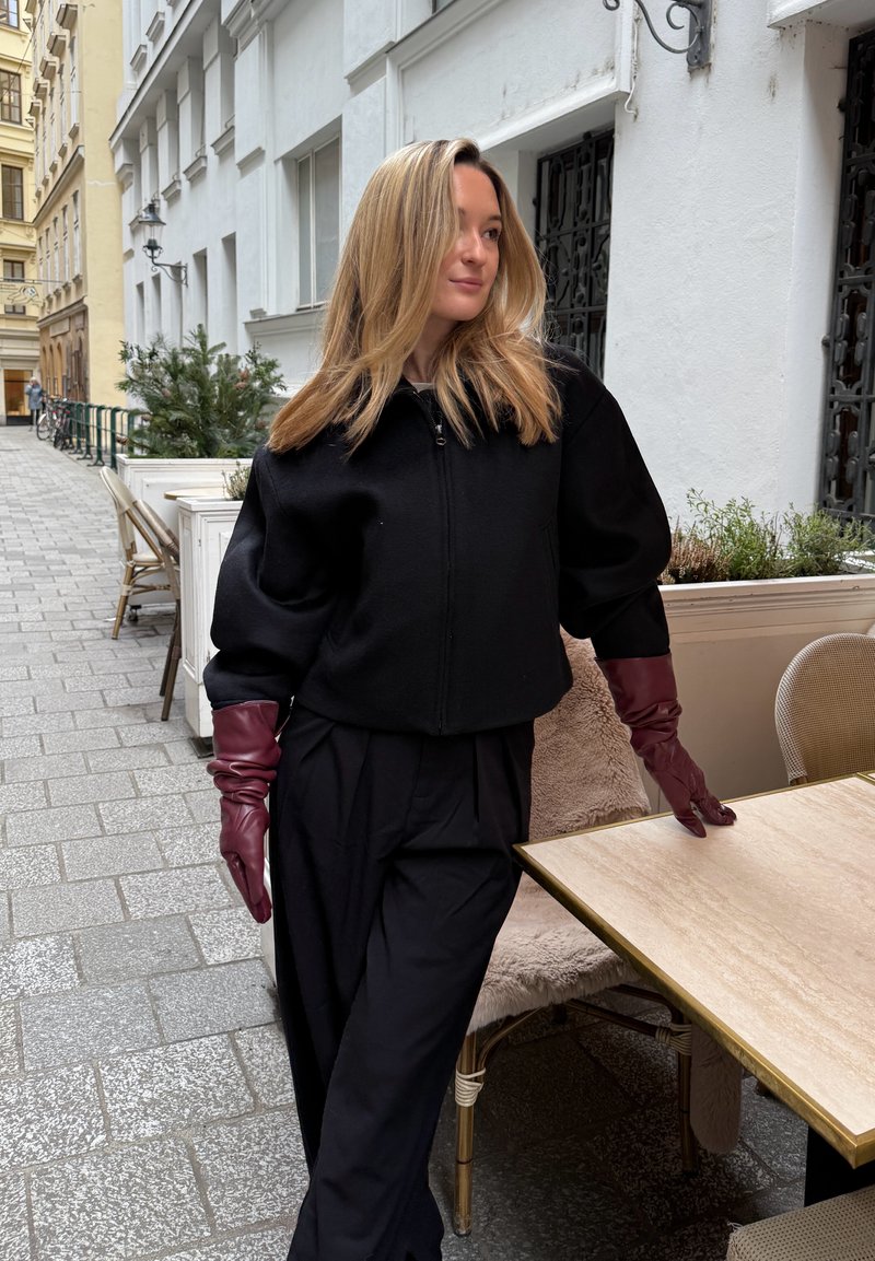 Zwarte kort gesneden jas met ritssluiting, pofmouwen, gecombineerd met een zwarte broek met hoge taille en bordeauxrode leren handschoenen, gesitueerd in een buiten café.