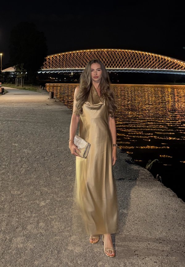 Une femme en robe dorée tient une pochette argentée au bord d’une rivière, la nuit, avec un pont en arc illuminé derrière elle.