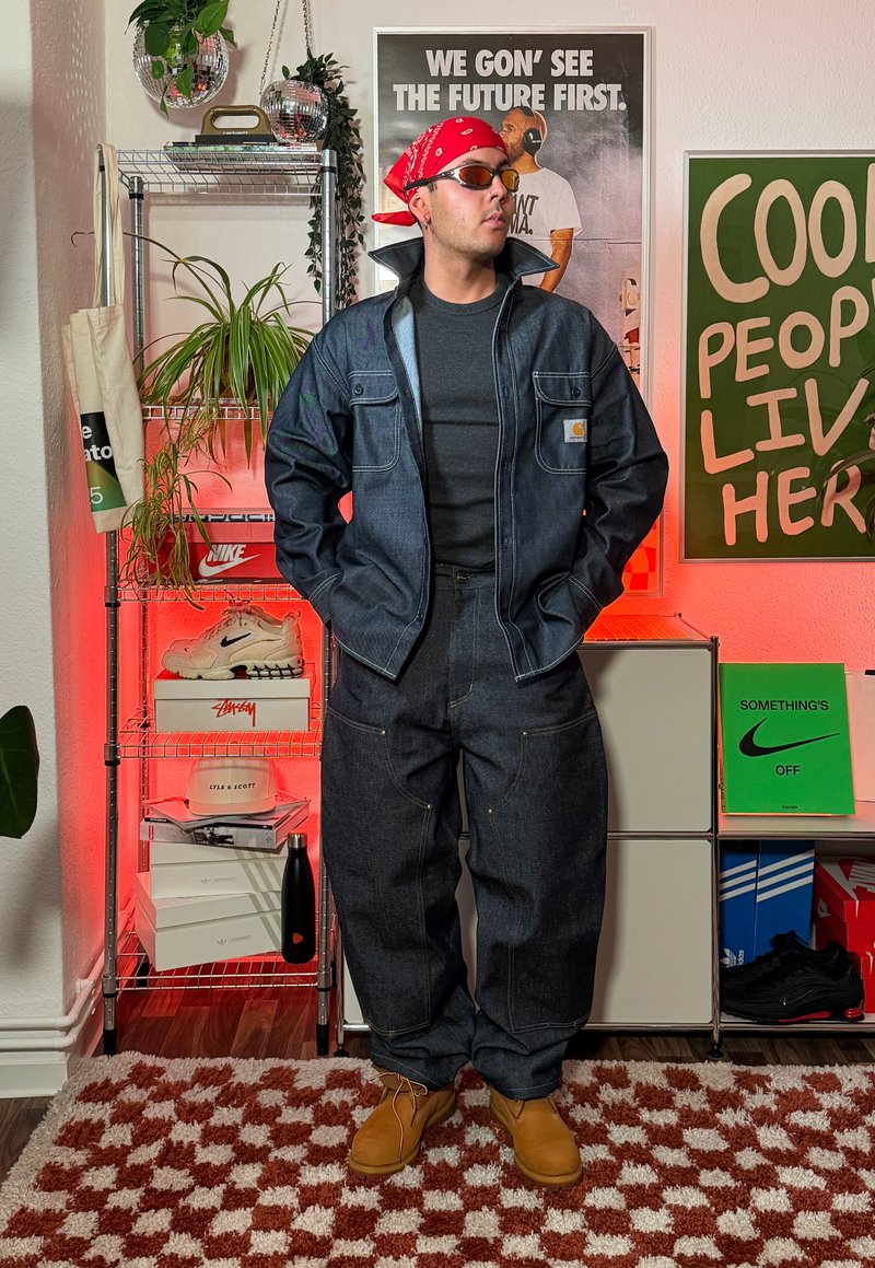 Veste en denim avec coutures apparentes et poches, associée à un pantalon en denim ample. T-shirt noir en dessous, chaussures marron, et bandana rouge.