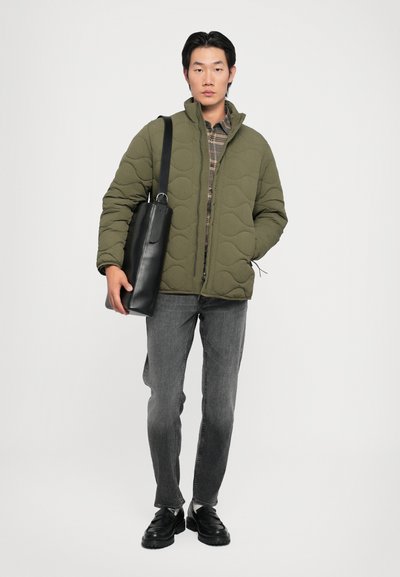 Chaqueta verde acolchada con cuello alto, camisa de cuadros gris debajo, jeans grises y bolso de cuero negro al hombro junto con zapatos negros.