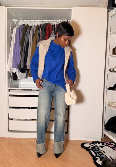 Mujer con suéter azul, chaleco de punto beige, vaqueros con puños y tacones negros sosteniendo un pequeño bolso beige en un vestidor.