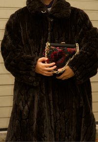 Manteau en fausse fourrure noire avec un motif texturé, fermeture à boutons, et un sac à main floral compact avec une bride chaîne dorée.