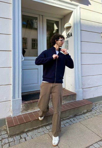 Joven con pelo rizado y gafas cerrándose una chaqueta azul marino mientras baja por la entrada del edificio número 36A.