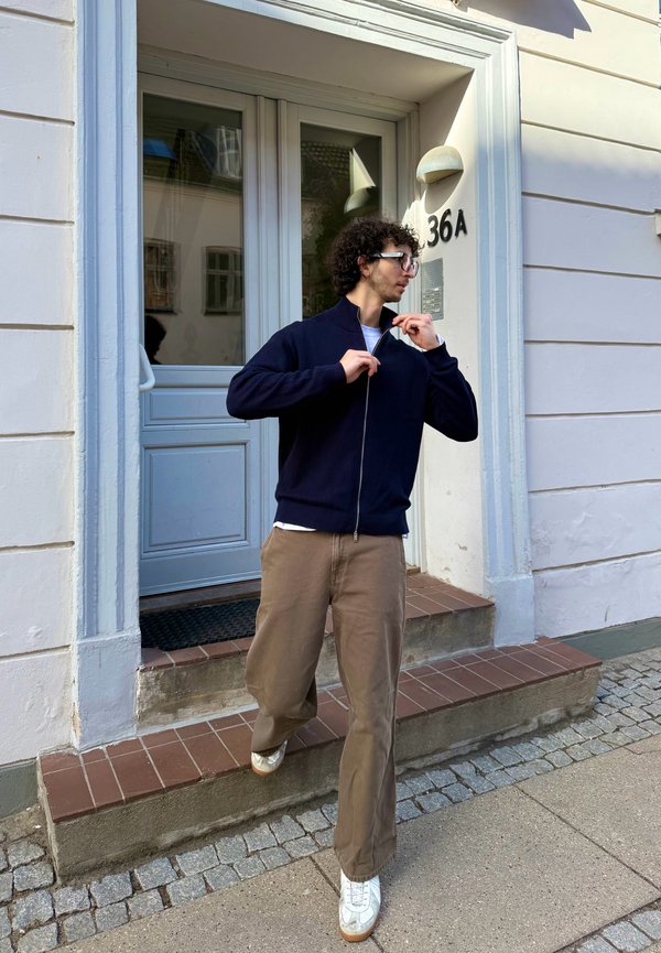 Junger Mann mit lockigem Haar und Brille zieht eine marineblaue Jacke zu, während er den Gebäudeeingang Nummer 36A hinuntersteigt.
