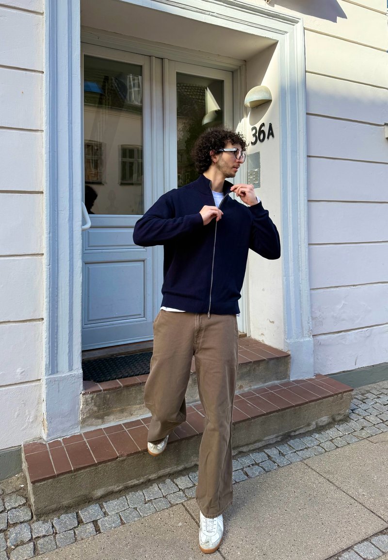 Jeune homme aux cheveux bouclés et lunettes en train de fermer sa veste bleu marine tout en descendant l'entrée du bâtiment numéro 36A.