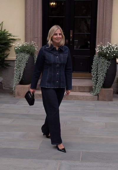 Chaqueta de mezclilla oscura con cierre de botones y bolsillos, combinada con pantalones anchos oscuros. Llevada con tacones negros de punta y un bolso negro acolchado.