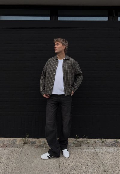 Camisa de cuadros en tonos oscuros y claros, camiseta blanca, jeans oscuros y zapatillas blancas con rayas negras en una calle contra una pared negra.