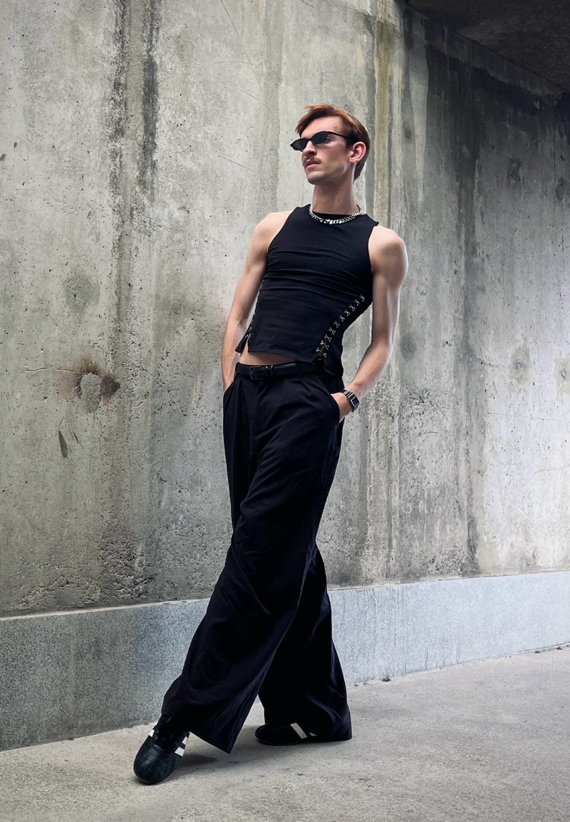 Jeune personne vêtue d'un haut noir sans manches, d'un pantalon large et de lunettes de soleil, appuyée contre un mur en béton avec les mains dans les poches.