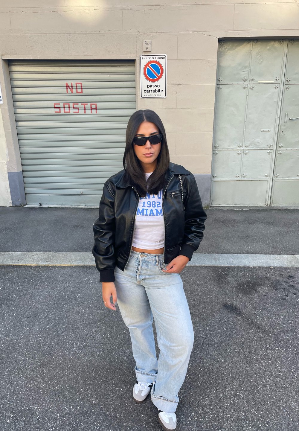 Giubbotto bomber in ecopelle nera sopra una maglietta bianca corta con scritte blu, jeans chiari a vita alta e sneaker grigie e bianche.