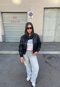 Giubbotto bomber in ecopelle nera sopra una maglietta bianca corta con scritte blu, jeans chiari a vita alta e sneaker grigie e bianche.