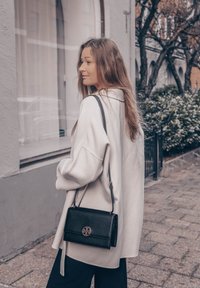 Svijetlo sivi oversized kaput s opuštenim krojem, uparen s malom crnom kožnom torbom preko ramena koja ima okrugli logo. Glatka tekstura, minimalistički dizajn.