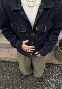 Osoba v tmavé džínové bundě přes bílý svetr, olivové cargo kalhoty, černé boty a stříbrné prsteny stojí na kamenných schodech a štěrkovém povrchu.