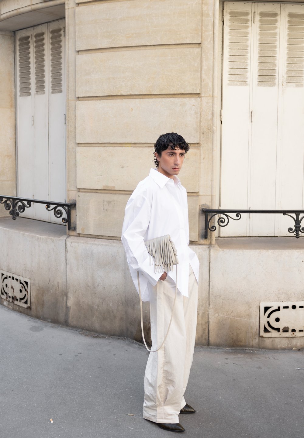 Camicia bianca oversize, pantaloni larghi color crema e una borsa leggera grigia con frange. La figura è appoggiata a una parete di pietra chiara e testurizzata.
