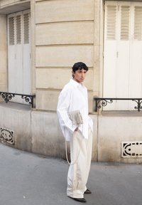 Camicia bianca oversize, pantaloni larghi color crema e una borsa leggera grigia con frange. La figura è appoggiata a una parete di pietra chiara e testurizzata.