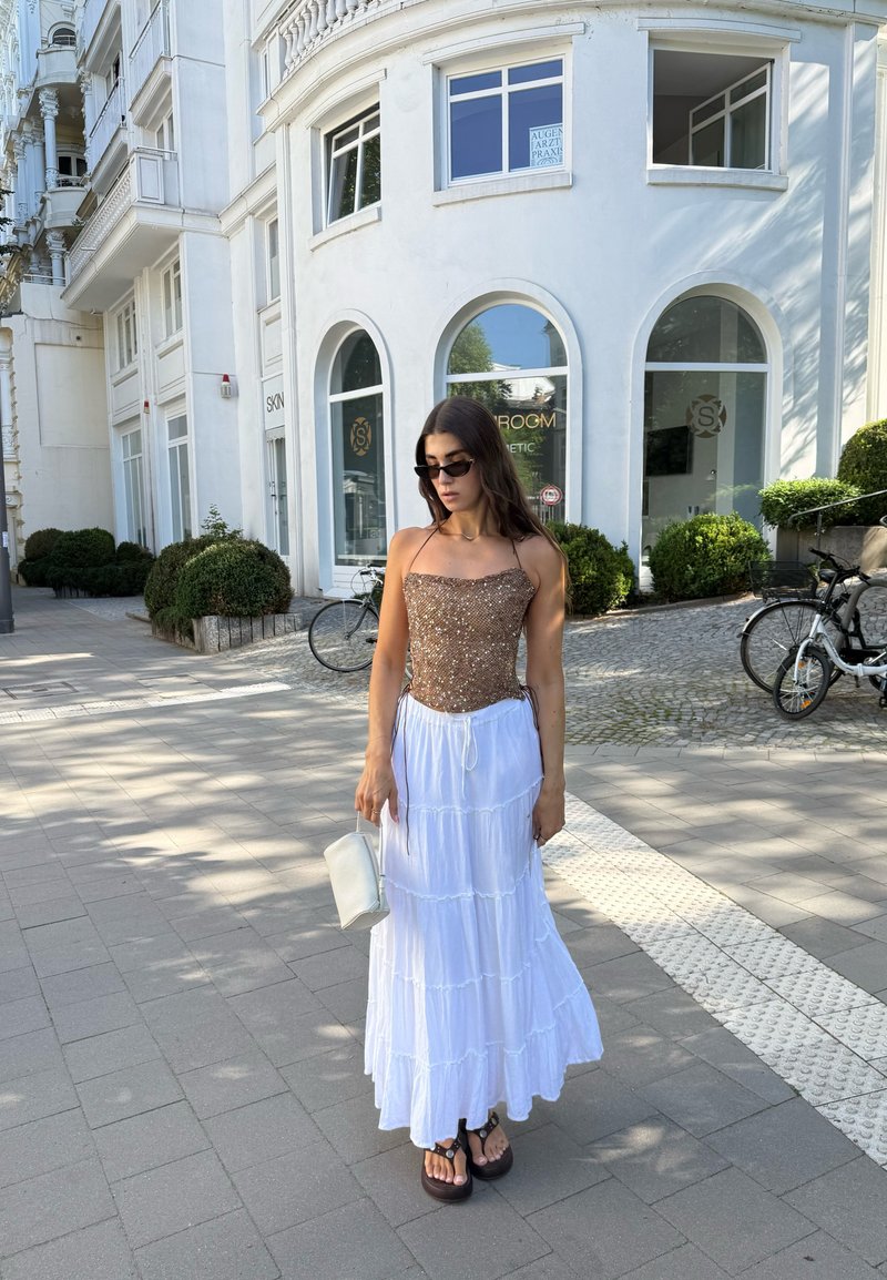 Femme portant des lunettes de soleil, un haut marron à paillettes, une jupe blanche à étages et des sandales, debout sur le trottoir près d'un bâtiment blanc et des bicyclettes.