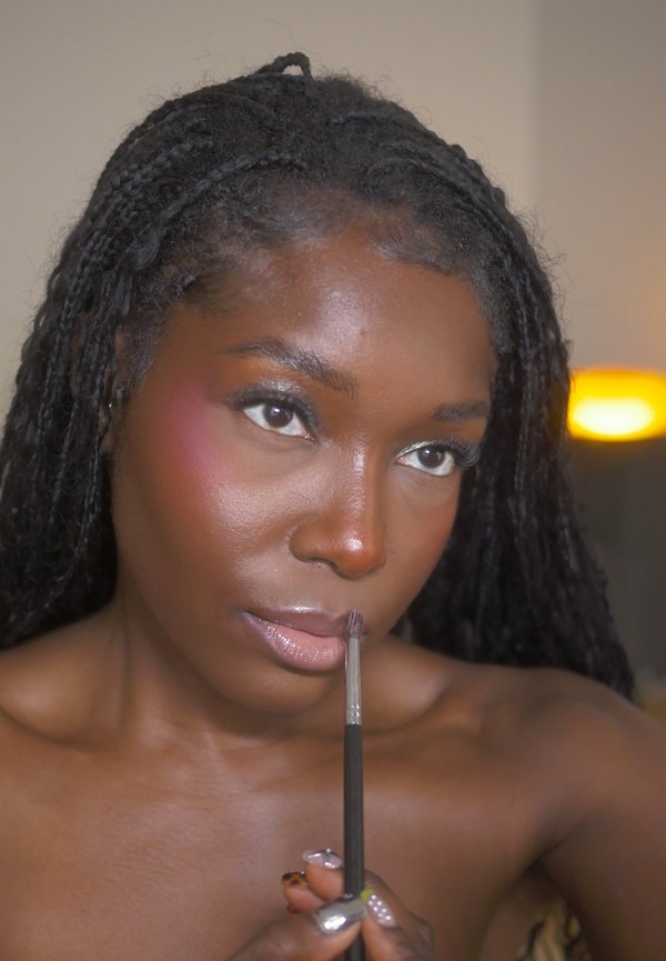 Femme aux cheveux tressés applique du maquillage sur ses lèvres avec un pinceau, en se concentrant sur ses lèvres, avec un arrière-plan doucement éclairé.