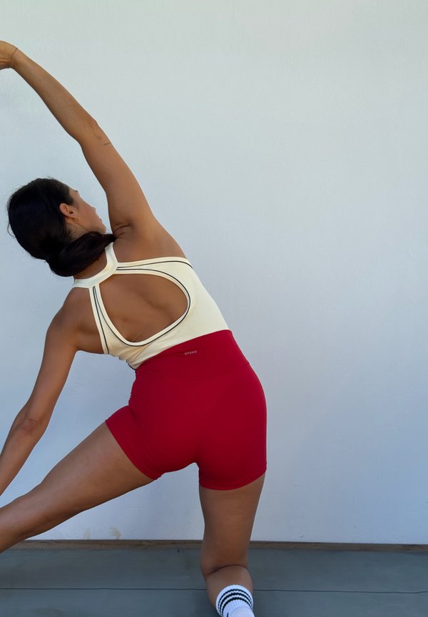 Femme en short rouge et haut blanc s'étirant latéralement avec un bras levé contre un mur clair uni.
