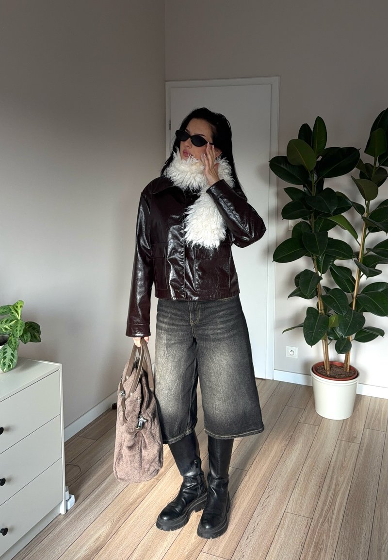 Bruine leren cropped jas met een witte nepbont kraag, gecombineerd met wijde, donkere jeans en zwarte veterboots; houdt een grijze tas vast.