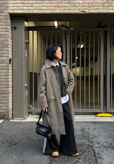 Persona con abrigo largo a cuadros, pantalones negros con rayas blancas, zapatos marrón claro, sosteniendo un bolso negro, de pie junto a una puerta metálica cerrada en un entorno urbano.