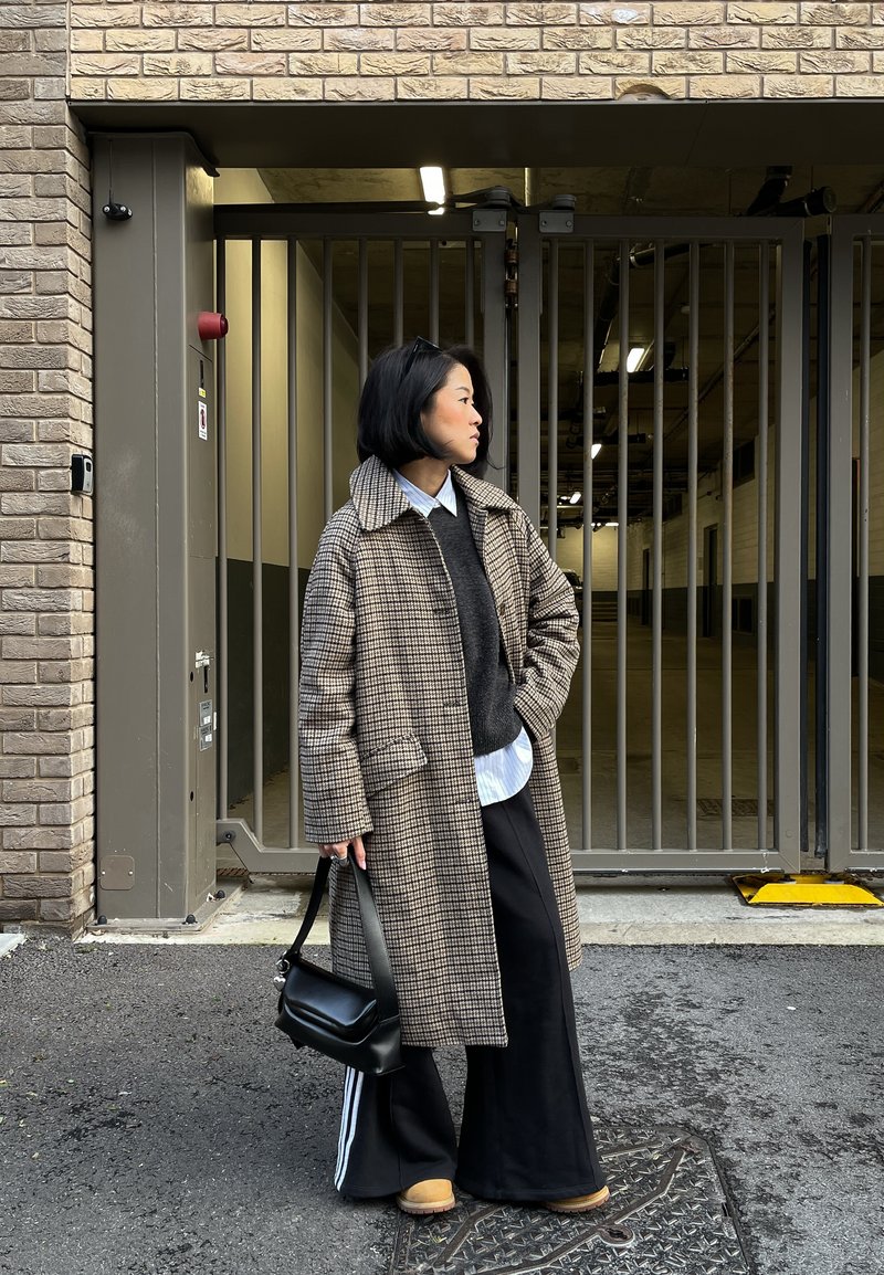 Personne portant un manteau long à carreaux, pantalon noir à rayures blanches, chaussures beige, tenant un sac à main noir, debout près d'une porte métallique fermée dans un environnement urbain.